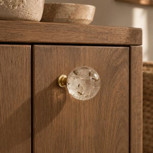 May include: A spherical clear resin cabinet knob featuring embedded dandelion seeds, attached to a circular brass base. The knob is mounted on a vertical wooden cabinet door with a visible grain pattern, showcasing a natural brown finish.