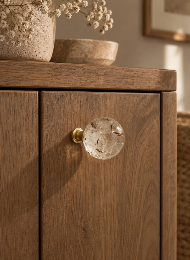May include: A spherical clear resin cabinet knob featuring embedded dandelion seeds, attached to a circular brass base. The knob is mounted on a vertical wooden cabinet door with a visible grain pattern, showcasing a natural brown finish.