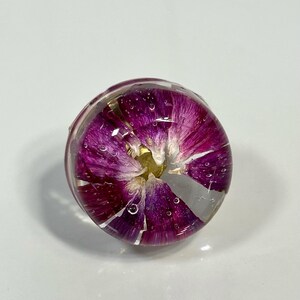 May include: A circular resin ring featuring a vibrant purple flower petal arrangement encased within clear, polished material. The design displays deep magenta and violet hues with a small yellow center, showing tiny air bubbles throughout the dome.