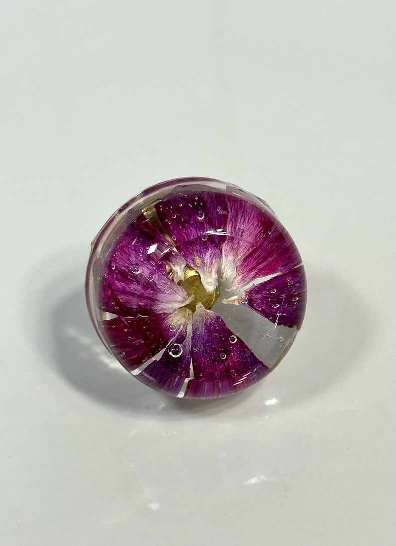 May include: A circular resin ring featuring a vibrant purple flower petal arrangement encased within clear, polished material. The design displays deep magenta and violet hues with a small yellow center, showing tiny air bubbles throughout the dome.