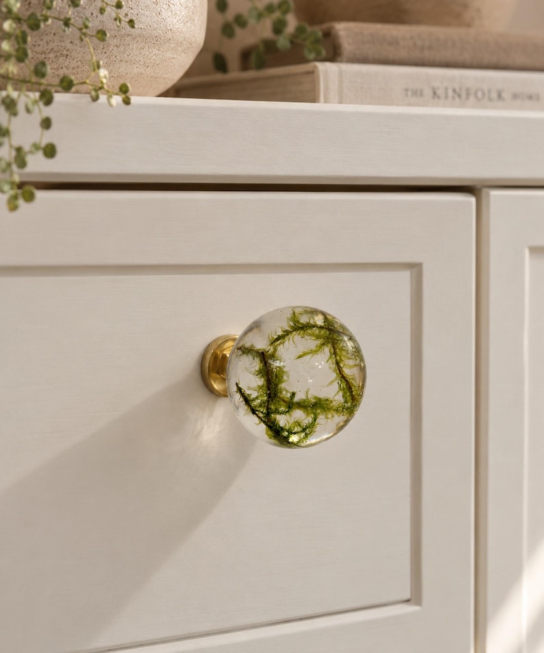 May include: A spherical clear resin cabinet knob featuring embedded green moss, attached to a circular brass-toned metal base. The knob is mounted on a flat, off-white cabinet drawer front with a recessed panel design.