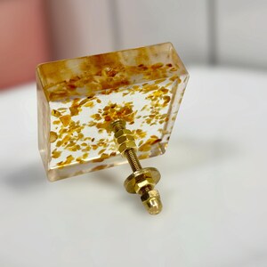 Op de afbeelding: Een vierkante meubelknop van heldere hars met ingebedde goudkleurige metaalvlokken. De knop is voorzien van een centrale goudkleurige metalen bout met een schroefdraad, een platte ring en een zeskantmoer aan de achterzijde voor montage.