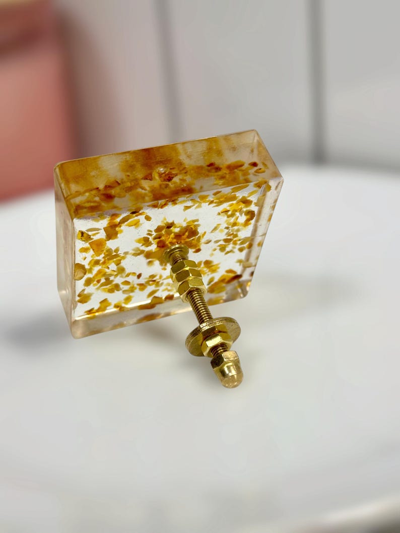 Op de afbeelding: Een vierkante meubelknop van heldere hars met ingebedde goudkleurige metaalvlokken. De knop is voorzien van een centrale goudkleurige metalen bout met een schroefdraad, een platte ring en een zeskantmoer aan de achterzijde voor montage.