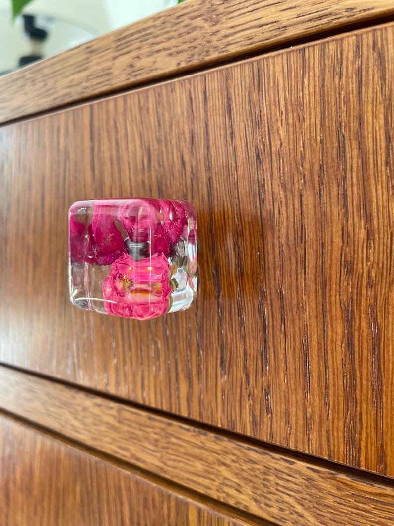 May include: A square-shaped drawer knob made of clear resin, featuring embedded bright pink and magenta flower petals throughout the interior. The knob is attached to a wooden drawer front with a visible wood grain pattern and horizontal lines.