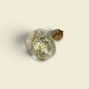 K&ouml;nnte beinhalten: Ein runder M&ouml;belknopf aus klarem Harz mit eingebetteten getrockneten wei&szlig;en Bl&uuml;ten und gr&uuml;nem Blattwerk. Der Knopf verf&uuml;gt &uuml;ber einen goldfarbenen Metallsockel mit einer Gewindeschraube an der R&uuml;ckseite zur Befestigung an M&ouml;beln.