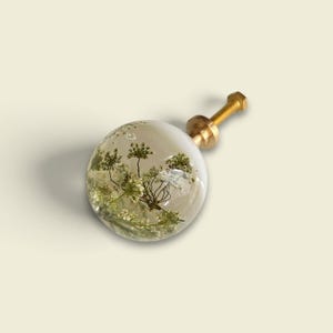 May include: A spherical cabinet knob made of clear resin containing preserved white and green botanical elements. The knob features a gold-toned metal mounting post with a threaded screw, designed for attachment to furniture or cabinetry doors.