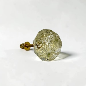 May include: A faceted, clear resin cabinet knob containing embedded dried white flowers and gold-colored glitter. The knob features a geometric, multi-sided shape and is attached to a brass-toned metal bolt with a matching washer and nut.