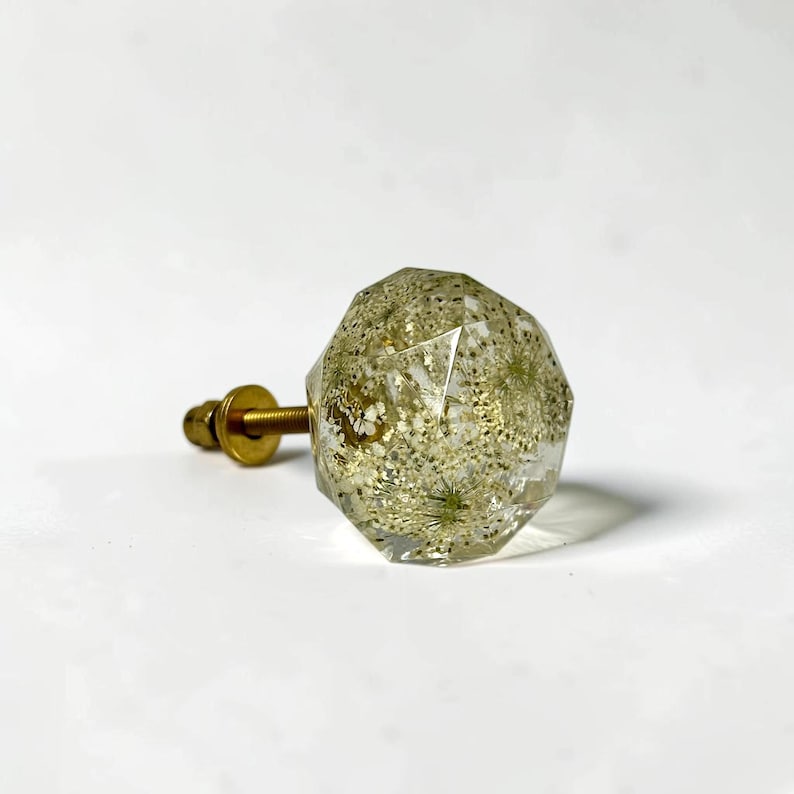 May include: A faceted, clear resin cabinet knob containing embedded dried white flowers and gold-colored glitter. The knob features a geometric, multi-sided shape and is attached to a brass-toned metal bolt with a matching washer and nut.