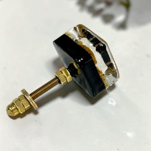 K&ouml;nnte beinhalten: Ein quadratischer M&ouml;belknopf mit einer transparenten Harzoberfl&auml;che, in die goldfarbene Flocken eingebettet sind, und einem soliden schwarzen Sockel. Der Knopf verf&uuml;gt &uuml;ber einen goldenen Metallbolzen mit zwei Muttern und einer Unterlegscheibe.