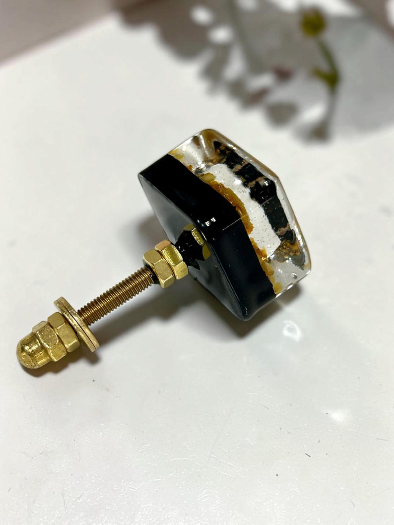 K&ouml;nnte beinhalten: Ein quadratischer M&ouml;belknopf mit einer transparenten Harzoberfl&auml;che, in die goldfarbene Flocken eingebettet sind, und einem soliden schwarzen Sockel. Der Knopf verf&uuml;gt &uuml;ber einen goldenen Metallbolzen mit zwei Muttern und einer Unterlegscheibe.