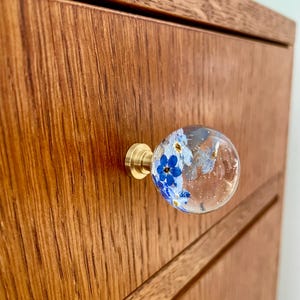 Op de afbeelding: Een bolvormige meubelknop met een heldere harskoepel waarin kleine blauwe vergeet-mij-nietjes en subtiele goudkleurige spikkels zijn verwerkt, bevestigd op een ronde basis van geborsteld messing op een lichte houten lade.
