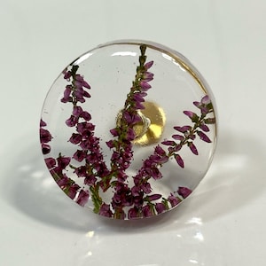 May include: A circular clear resin knob featuring embedded purple heather flowers. The knob has a smooth, transparent finish and a gold-toned metal attachment point visible in the center, showcasing the natural botanical details within the resin.