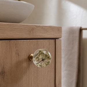 May include: A spherical cabinet knob made of clear resin containing a dried white flower, attached to a circular brass base. The knob is mounted on a light-toned wooden cabinet door with a visible grain pattern, featuring a smooth, polished finish.