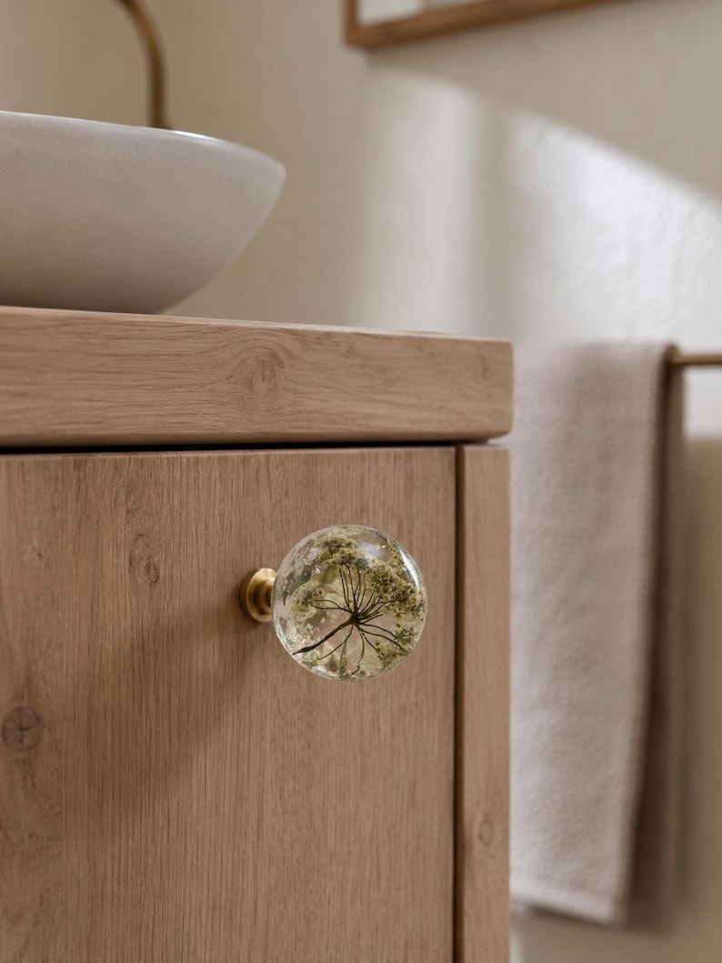 May include: A spherical cabinet knob made of clear resin containing a dried white flower, attached to a circular brass base. The knob is mounted on a light-toned wooden cabinet door with a visible grain pattern, featuring a smooth, polished finish.