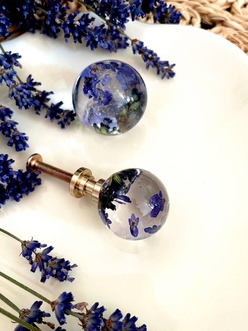 May include: Two spherical drawer knobs made of clear resin, each containing dried purple lavender flowers. One knob is attached to a metallic gold-toned screw base, while the other is detached. The knobs are surrounded by sprigs of fresh lavender.