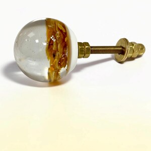 K&ouml;nnte beinhalten: Ein kugelf&ouml;rmiger M&ouml;belknopf mit einer klaren, transparenten Harzh&auml;lfte und einem strukturierten, bernsteinfarbenen Innenteil. Der Knopf ist an einem messingfarbenen Gewindebolzen aus Metall mit passender Unterlegscheibe und Mutter befestigt.