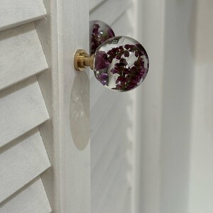K&ouml;nnte beinhalten: Ein kugelf&ouml;rmiger Schrankknauf aus klarem Harz mit eingebetteten violetten Trockenblumen. Der Knauf ist an einem messingfarbenen Metallsockel befestigt und an einer wei&szlig;en Lamellent&uuml;r montiert. Das Design weist eine glatte, abgerundete Oberfl&auml;che auf.