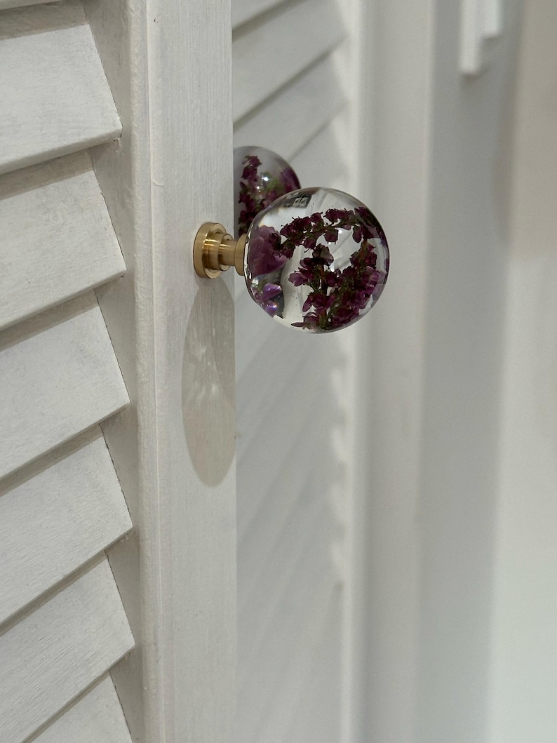 K&ouml;nnte beinhalten: Ein kugelf&ouml;rmiger Schrankknauf aus klarem Harz mit eingebetteten violetten Trockenblumen. Der Knauf ist an einem messingfarbenen Metallsockel befestigt und an einer wei&szlig;en Lamellent&uuml;r montiert. Das Design weist eine glatte, abgerundete Oberfl&auml;che auf.
