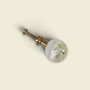 May include: A spherical cabinet knob featuring a clear resin dome with embedded white dried flowers. The knob is attached to a gold-toned metal base and a threaded screw, providing a functional hardware piece for furniture or cabinetry doors.