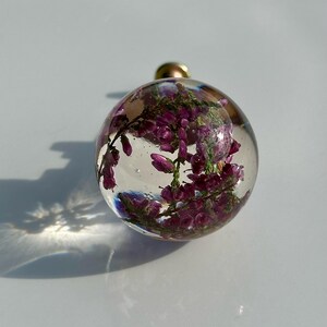 K&ouml;nnte beinhalten: Ein kugelf&ouml;rmiger Knauf aus klarem Harz mit eingebetteten violetten Heidekrautbl&uuml;ten. Die transparente Kugel hat eine glatte, polierte Oberfl&auml;che und ist an einem kleinen Metallsockel befestigt, was eine botanische Darstellung in runder Form ergibt.