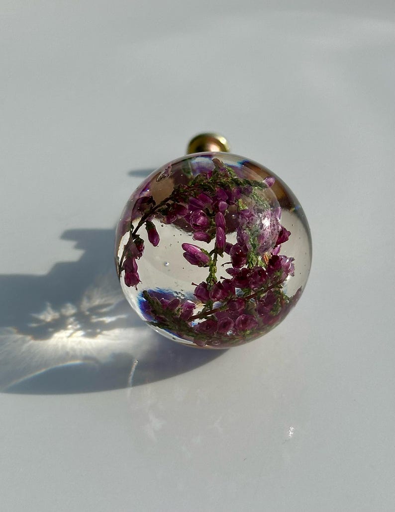 K&ouml;nnte beinhalten: Ein kugelf&ouml;rmiger Knauf aus klarem Harz mit eingebetteten violetten Heidekrautbl&uuml;ten. Die transparente Kugel hat eine glatte, polierte Oberfl&auml;che und ist an einem kleinen Metallsockel befestigt, was eine botanische Darstellung in runder Form ergibt.