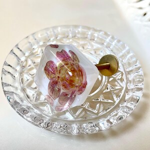 May include: A hexagonal cabinet knob made of clear resin containing a pressed pink hydrangea flower. The knob features a gold-toned metal screw and washer attached to the back, resting inside a small, round, clear glass dish with a faceted edge.