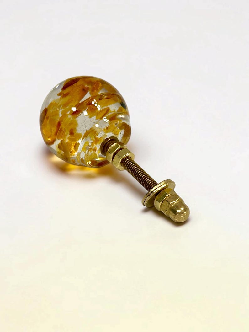 Puede incluir: Un pomo esf&eacute;rico para muebles hecho de resina transparente con escamas de color &aacute;mbar incrustadas. El pomo cuenta con un perno, una arandela y una tuerca de metal dorado en la parte posterior, formando un mecanismo de fijaci&oacute;n funcional.