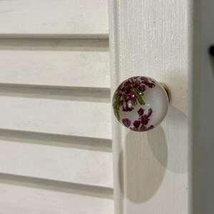 May include: A round cabinet knob featuring a clear, spherical resin body with embedded purple heather flowers and green foliage. The knob is attached to a gold-toned metal base and is mounted on a white, louvered wooden cabinet door panel.