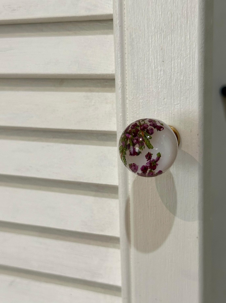 May include: A round cabinet knob featuring a clear, spherical resin body with embedded purple heather flowers and green foliage. The knob is attached to a gold-toned metal base and is mounted on a white, louvered wooden cabinet door panel.