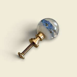 K&ouml;nnte beinhalten: Ein kugelf&ouml;rmiger M&ouml;belknopf mit einer klaren Harzkuppel und eingebetteten blauen Blumen, befestigt an einem goldfarbenen Metallsockel mit Gewindeschraube. Der Knopf hat eine glatte, abgerundete Oberfl&auml;che und einen metallischen Schaft zur Montage.