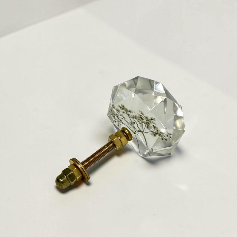 May include: A clear, faceted, diamond-shaped cabinet knob made of resin, featuring an embedded dried white flower inside. The knob is attached to a threaded brass-colored metal bolt with two matching hexagonal nuts and a flat washer.