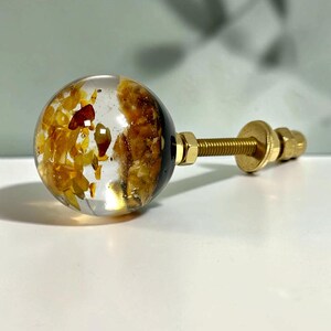 May include: A spherical cabinet knob featuring a clear resin body embedded with amber-colored stone fragments. The knob is attached to a gold-toned metal bolt with a matching washer and nut, providing a secure mounting mechanism for furniture.