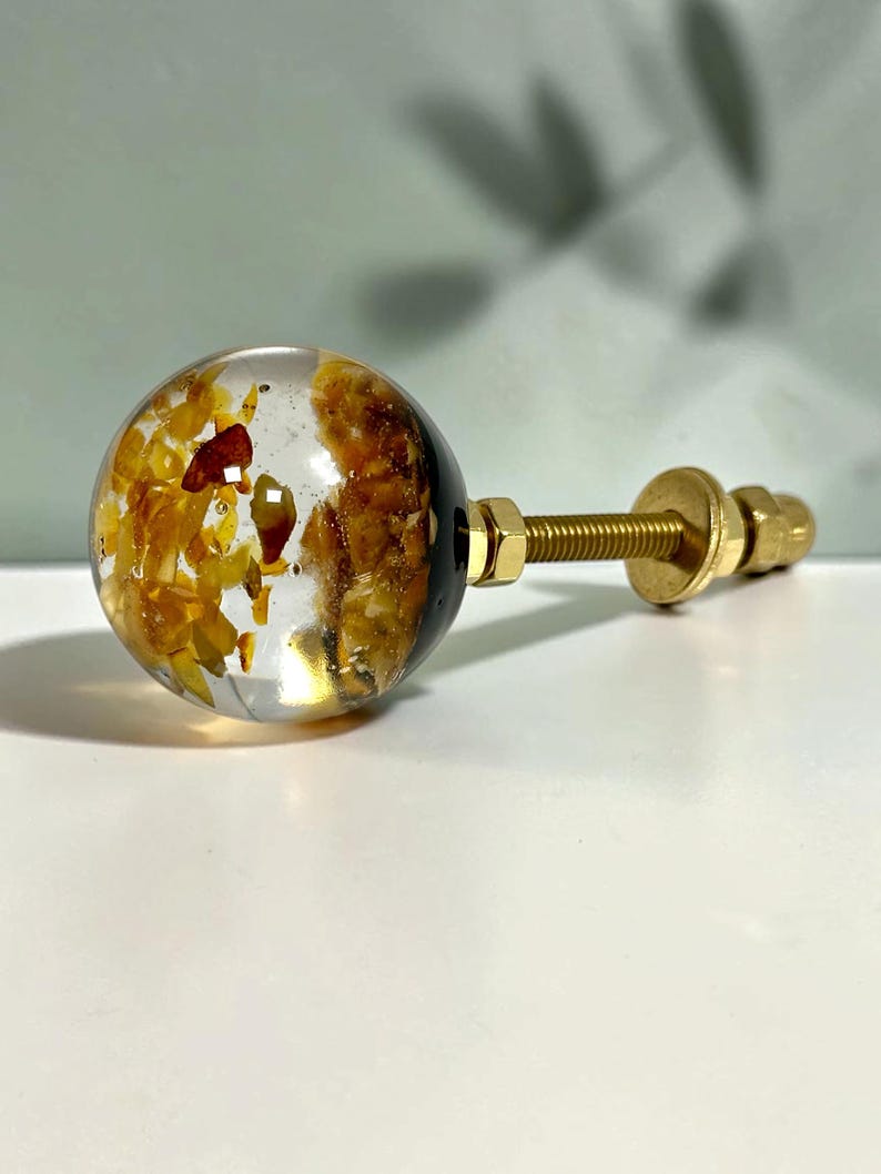 May include: A spherical cabinet knob featuring a clear resin body embedded with amber-colored stone fragments. The knob is attached to a gold-toned metal bolt with a matching washer and nut, providing a secure mounting mechanism for furniture.