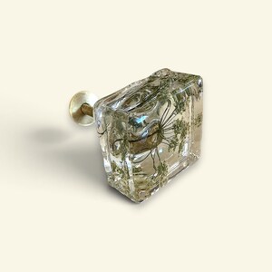 Puede incluir: Un tirador de mueble cuadrado hecho de resina transparente con una flor blanca prensada en su interior. El tirador cuenta con una base circular de tono dorado y un tornillo met&aacute;lico roscado que sobresale por detr&aacute;s para su fijaci&oacute;n.