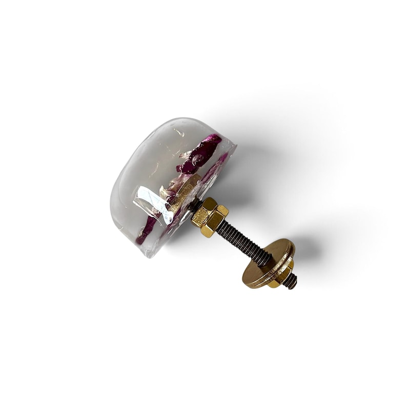 May include: A clear, semi-spherical resin cabinet knob featuring embedded dark purple flower petals. The knob includes a gold-toned metal bolt, a hexagonal nut, and a circular washer attached to the back for mounting purposes.