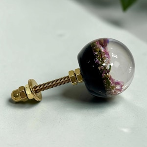 Pode incluir: Um puxador esf&eacute;rico para m&oacute;veis com a metade superior em resina transparente e a metade inferior preta. Dentro da parte clara, s&atilde;o vis&iacute;veis pequenas flores secas cor-de-rosa e verdes. O puxador tem um parafuso met&aacute;lico dourado com duas porcas.