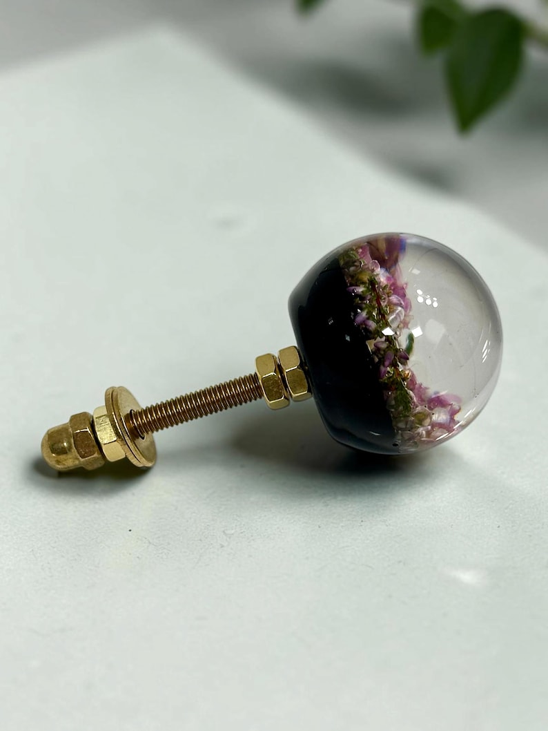 Pode incluir: Um puxador esf&eacute;rico para m&oacute;veis com a metade superior em resina transparente e a metade inferior preta. Dentro da parte clara, s&atilde;o vis&iacute;veis pequenas flores secas cor-de-rosa e verdes. O puxador tem um parafuso met&aacute;lico dourado com duas porcas.