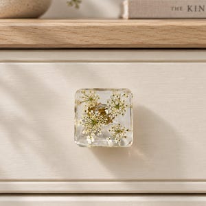 Puede incluir: Un tirador de mueble cuadrado hecho de resina transparente, con flores blancas prensadas incrustadas en su interior. El tirador tiene una base de metal dorado y está fijado a un frente de cajón de madera color crema con molduras.