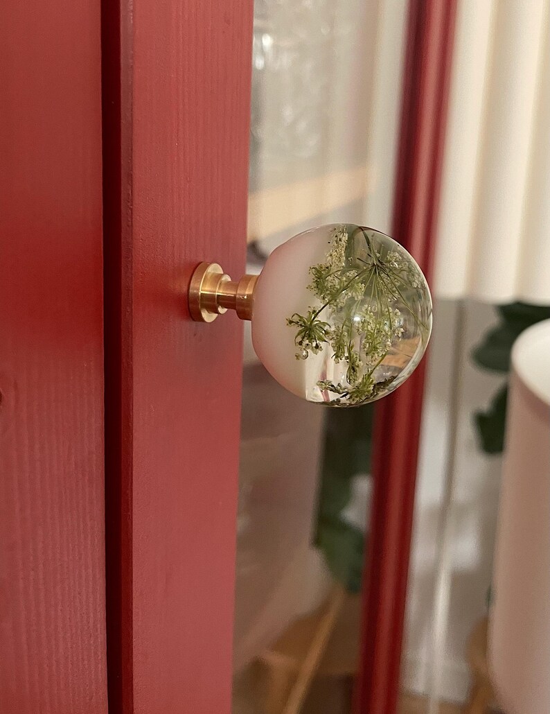 May include: A spherical cabinet knob featuring a clear resin dome with an embedded white pressed flower, attached to a cylindrical brass base. The knob is mounted on a red wooden surface, showcasing a smooth, rounded profile and metallic hardware.