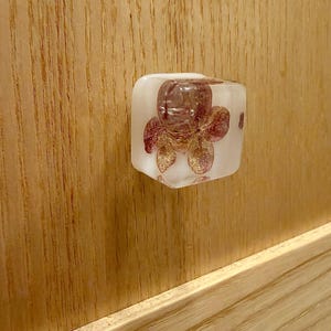 May include: A square-shaped cabinet knob made of clear resin with a translucent white base. Embedded inside the resin is a dried, pressed flower with delicate pink and purple petals, visible through the front face of the knob.