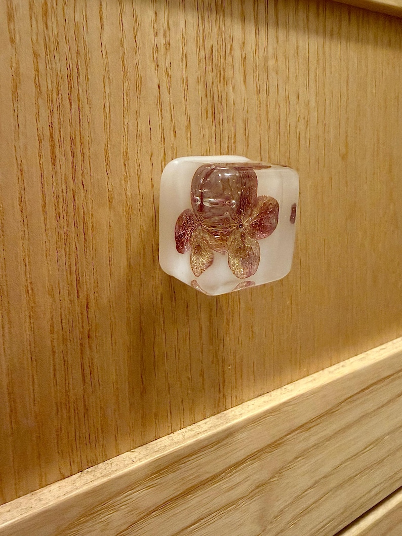 May include: A square-shaped cabinet knob made of clear resin with a translucent white base. Embedded inside the resin is a dried, pressed flower with delicate pink and purple petals, visible through the front face of the knob.