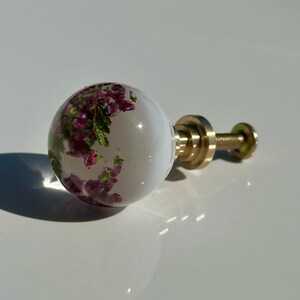 May include: A spherical cabinet knob featuring a clear resin body with embedded purple and green heather flowers. The knob is attached to a gold-toned metal base and a threaded screw, providing a functional hardware piece for furniture.