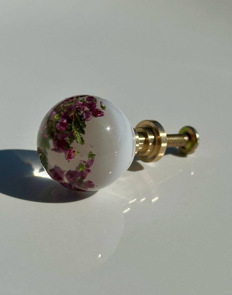May include: A spherical cabinet knob featuring a clear resin body with embedded purple and green heather flowers. The knob is attached to a gold-toned metal base and a threaded screw, providing a functional hardware piece for furniture.