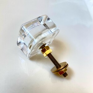 May include: A clear, faceted glass cabinet knob with a square-shaped head. The hardware includes a threaded brass-toned metal bolt, two hexagonal nuts, and a flat circular washer, all attached to the back of the transparent glass component.