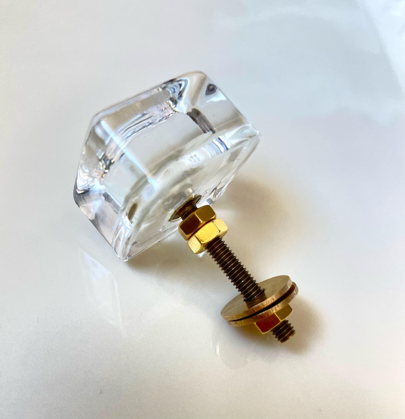 May include: A clear, faceted glass cabinet knob with a square-shaped head. The hardware includes a threaded brass-toned metal bolt, two hexagonal nuts, and a flat circular washer, all attached to the back of the transparent glass component.