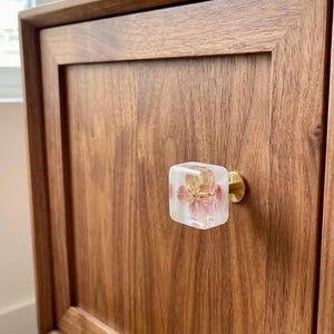 May include: A square cabinet knob featuring a clear resin block with an embedded pink and yellow flower, attached to a circular brass-toned metal base, mounted on a flat-panel wooden cabinet door with a visible natural wood grain pattern.