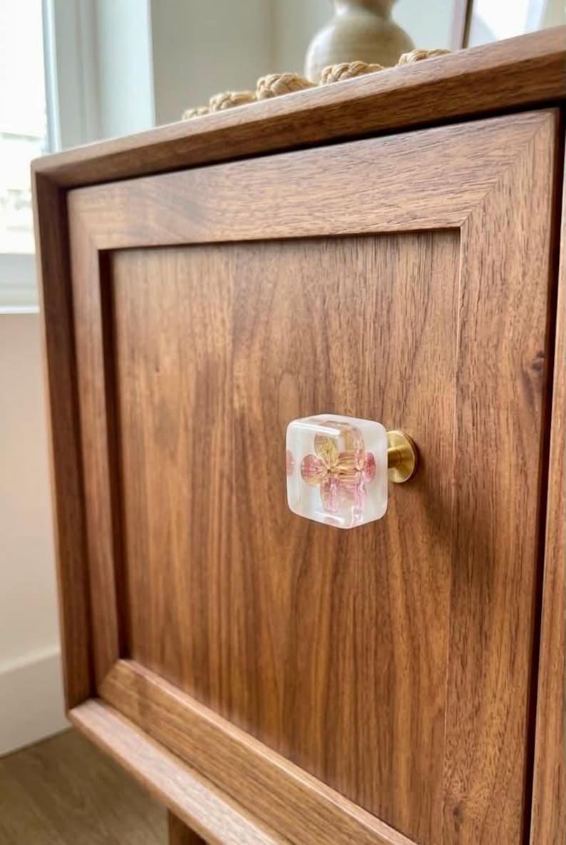 May include: A square cabinet knob featuring a clear resin block with an embedded pink and yellow flower, attached to a circular brass-toned metal base, mounted on a flat-panel wooden cabinet door with a visible natural wood grain pattern.