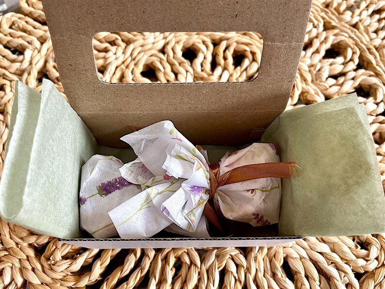 Puede incluir: Una peque&ntilde;a caja de cart&oacute;n marr&oacute;n con una abertura de asa, forrada con papel de seda verde claro. Dentro, un paquete envuelto en papel de seda blanco con estampado de flores de lavanda, atado con una cinta marr&oacute;n fina.