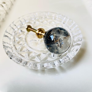 Puede incluir: Un pomo esf&eacute;rico para muebles con una c&uacute;pula de resina transparente que encierra semillas de diente de le&oacute;n blancas sobre un fondo oscuro, unido a una base de metal dorado con tornillo roscado, sobre un peque&ntilde;o plato de cristal transparente con borde facetado.