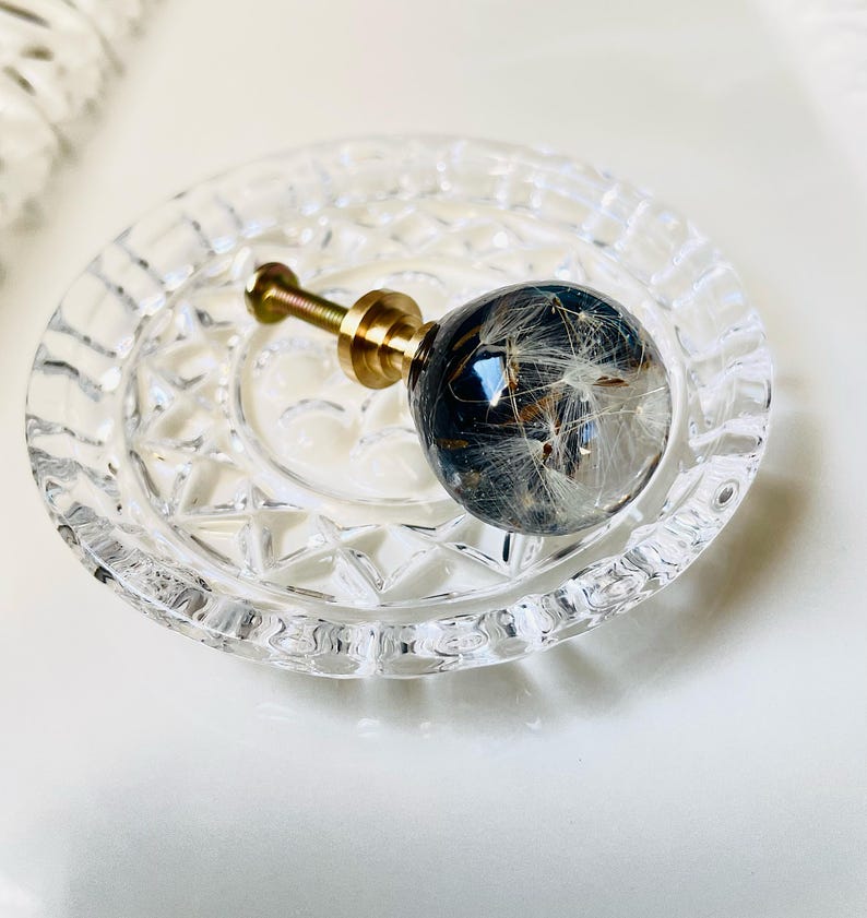 Puede incluir: Un pomo esf&eacute;rico para muebles con una c&uacute;pula de resina transparente que encierra semillas de diente de le&oacute;n blancas sobre un fondo oscuro, unido a una base de metal dorado con tornillo roscado, sobre un peque&ntilde;o plato de cristal transparente con borde facetado.